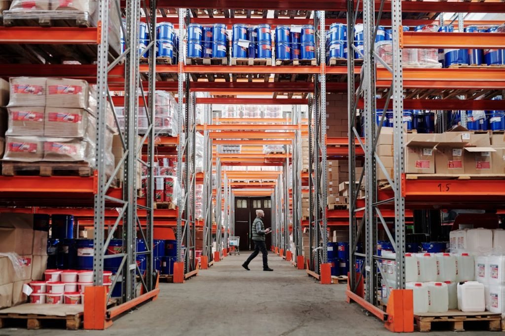man-walking-in-warehouse-4483775 A man walking through a large industrial warehouse with stacked shelves filled with goods and products.